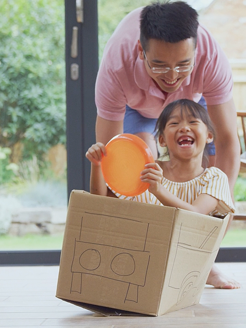 Dad pushing Daughter in Cardboard Box on Hardwood Floors